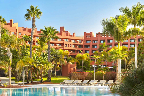 Groot zwembad voor het hotel Tivoli La Caleta, omringd door palmbomen, tuinen en ligbedden.